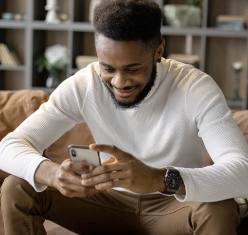 A black man sitting on a couch. He is smiling and is looking at the smartphone in his hands.