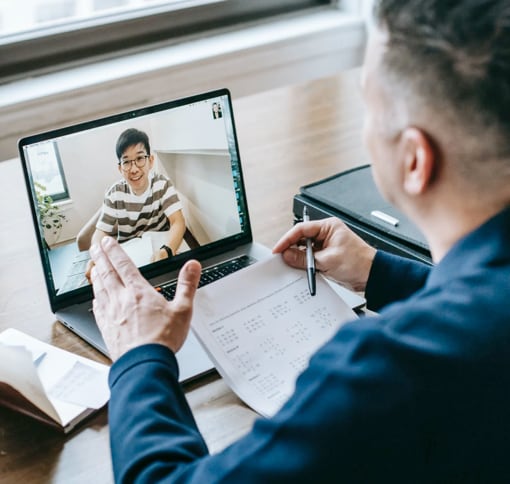 A white man sitting at a table looking at the screen of the laptop in front of him. He is in a video call with another man.