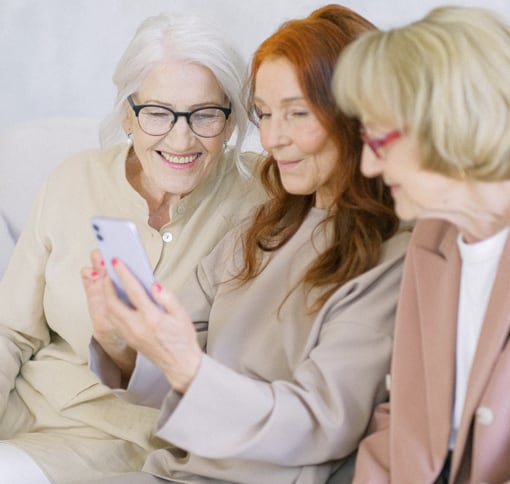 Three white women sitting next to each other. All three are looking at a smartphone that the middle woman is holding in her hands.